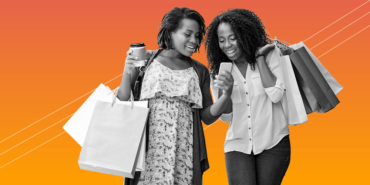 Two women standing and huddled together over a cell phone. Both are holding shopping bags.